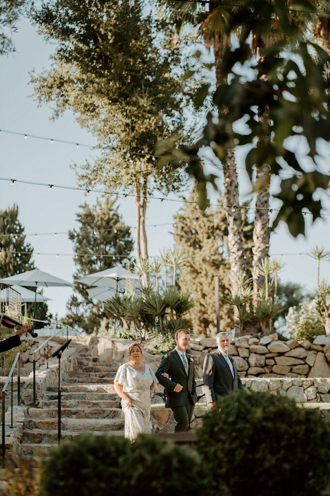 The groom and his parents walk into the outdoor wedding ceremony space at Mt Woodson Castle in California.  They walk down a set of stone steps in the California sun. 