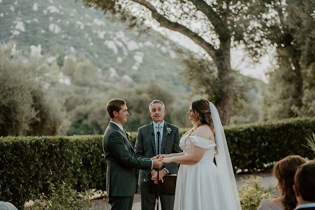 A bride and groom hold hands and exchange vows during their California wedding ceremony at Mt Woodson Castle.  Behind them is a line of bushes and large trees framed by a mountain. 