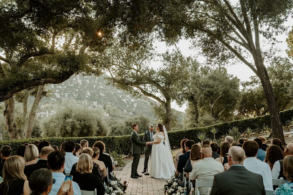 A bride and groom hold hands and exchange vows during their California wedding ceremony at Mt Woodson Castle.  Behind them is a line of bushes and large trees framed by a mountain.   The aisle is lined with white roses in large end cap installations. 