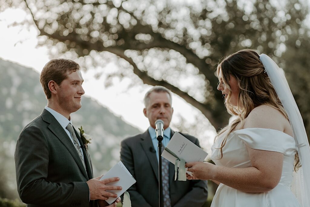 A bride and groom hold hands and exchange vows during their California wedding ceremony at Mt Woodson Castle.  Behind them is a line of bushes and large trees framed by a mountain. They are holding white vow books with sage green ribbons holding the booklet together.