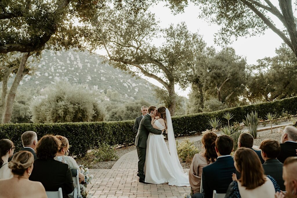 A bride and groom share a first married kiss during their California wedding ceremony at Mt Woodson Castle.  Behind them is a line of bushes and large trees framed by a mountain. 