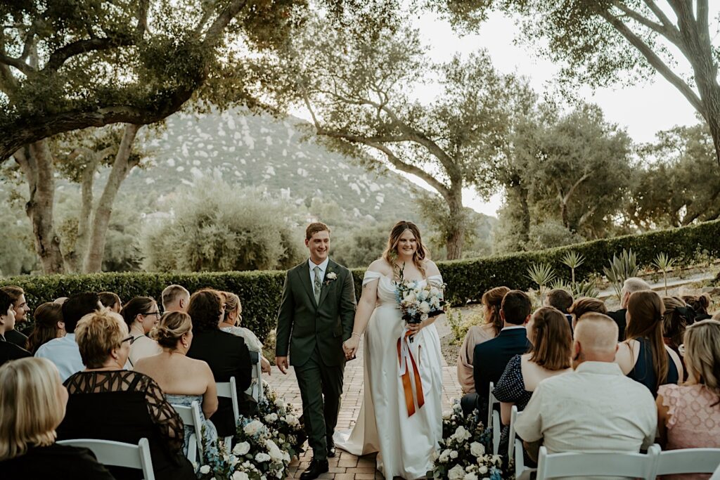 A bride and groom walk down the aisle holding hands during their California wedding ceremony at Mt Woodson Castle.  Behind them is a line of bushes and large trees framed by a mountain. 