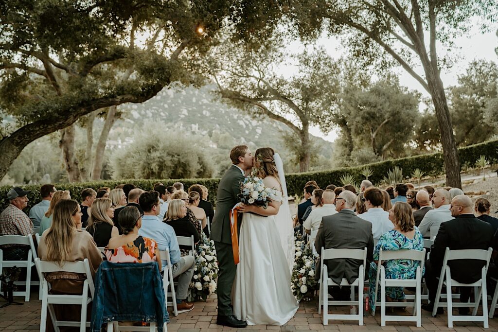 A bride and groom kiss while walking down the aisle after their California wedding ceremony at Mt Woodson Castle.  Behind them is a line of bushes and large trees framed by a mountain. 