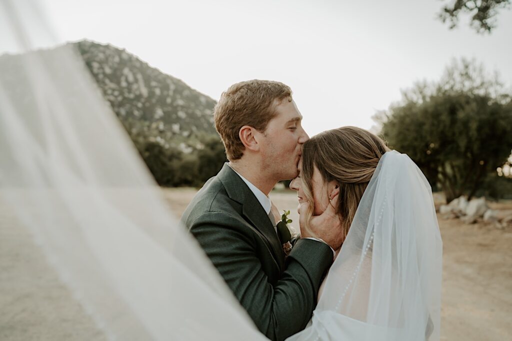 A groom kisses his bride on the forehead standing in the dessert at Mt Woodson Castle in California on their wedding day.  The brides long veil is stretched towards the camera.