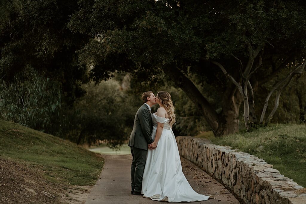 A bride and groom pause to kiss while walk through the lawn of Mt Woodson Castle.  They walk along a line of bricks on a path that is covered by trees.