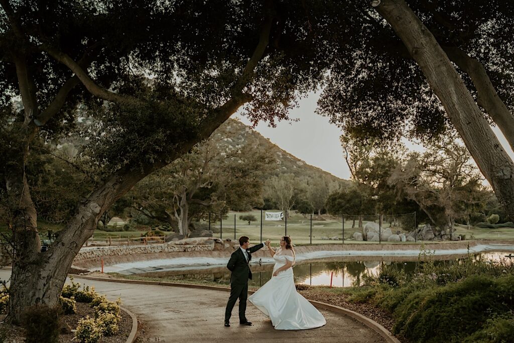 A bride and groom dance on a path at Mt Woodson Castle overlooking a lake on the golf course.