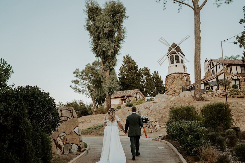 A bride and groom walk holding hands towards Mt Woodson Castle in California.  In the distance you can see the signature windmill.