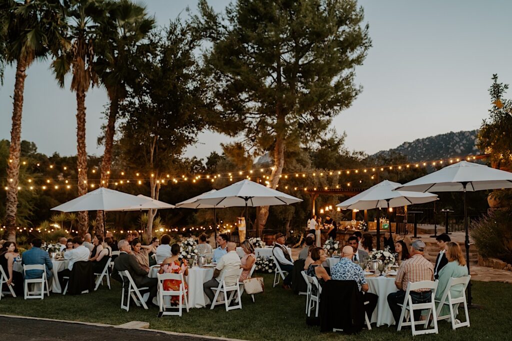 Guests sit and enjoy dinner on a lawn at Mt Woodson Castle in Ramona California during a bride and grooms destination wedding.