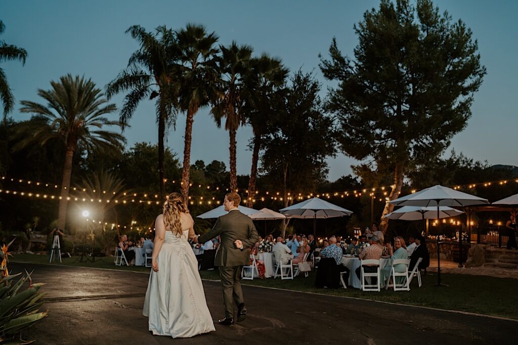 A bride and groom walk into their wedding reception an outdoor space covered in string lights at Mt Woodson Castle in California.  