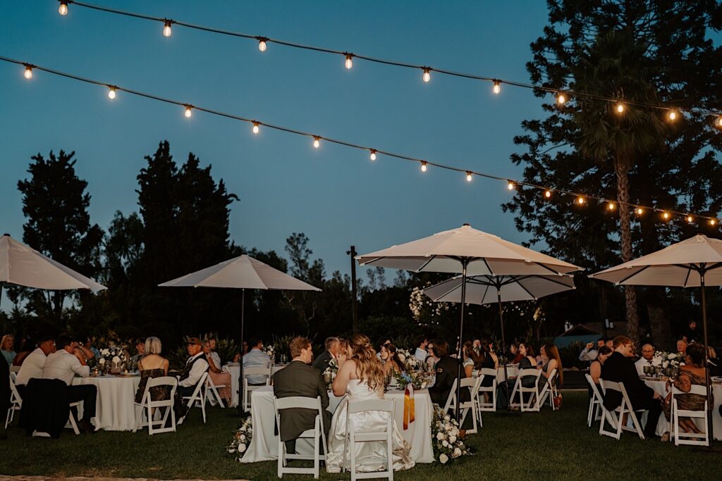 A bride and groom share a moment together at their sweetheart table at their wedding reception an outdoor space covered in string lights at Mt Woodson Castle in California.  