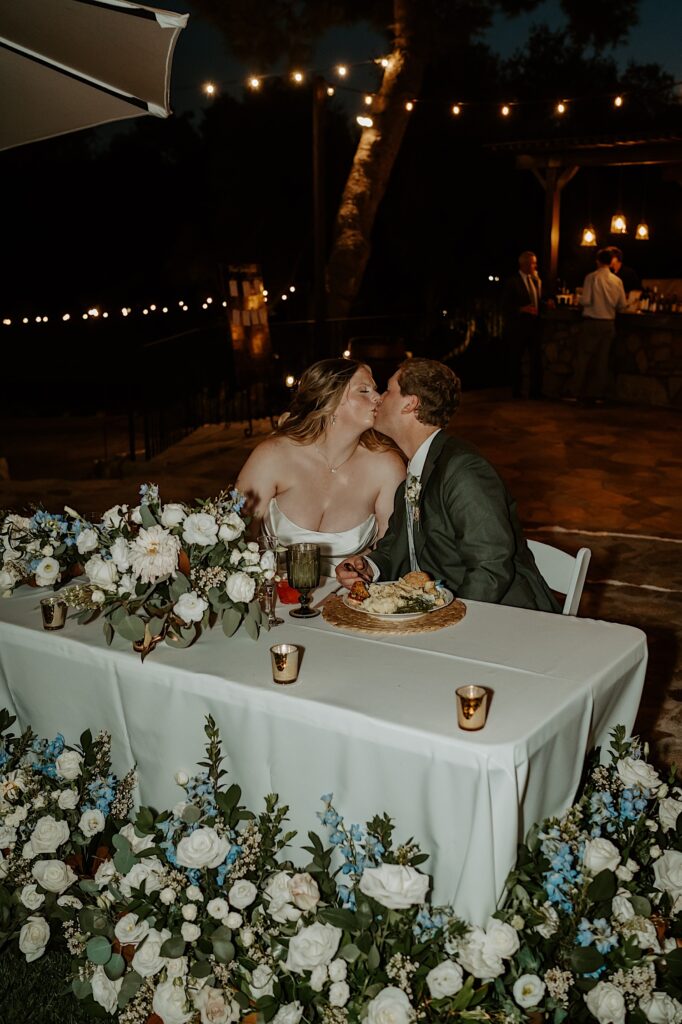 A bride and groom share a moment together at their sweetheart table at their wedding reception an outdoor space covered in string lights at Mt Woodson Castle in California.  