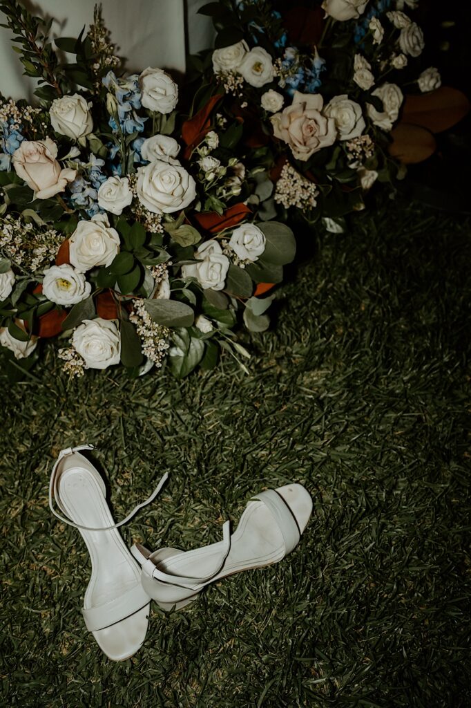 A brides wedding details photographed with flash photography.  Her wedding shoes sitting on the lawn in front of their orange and white and blue flowers.