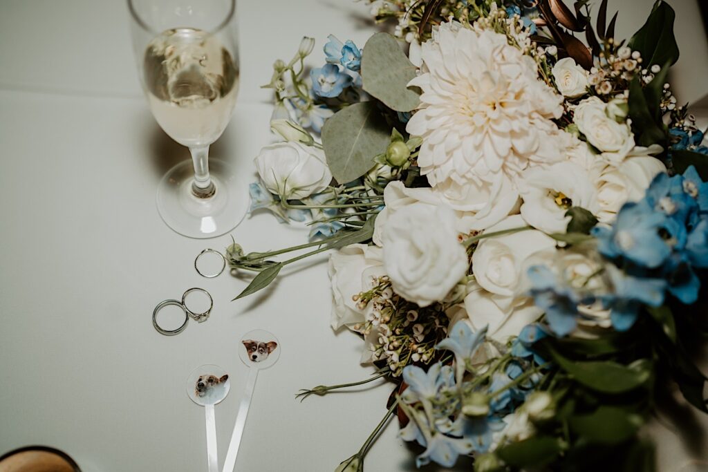 A brides and grooms wedding details photographed with flash photography.  Their wedding rings sit with drink stirrers with their pets faces on them, their rings sit scattered on the table and a glass of champagne sits with the bouquet.