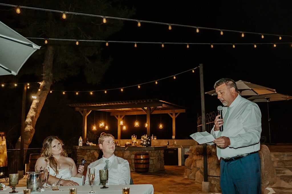 A guest gives a toast to the bride and groom during their outdoor wedding reception.  The reception is lit with string lights and the bride and groom smile at the guest giving the toast during their California Wedding.