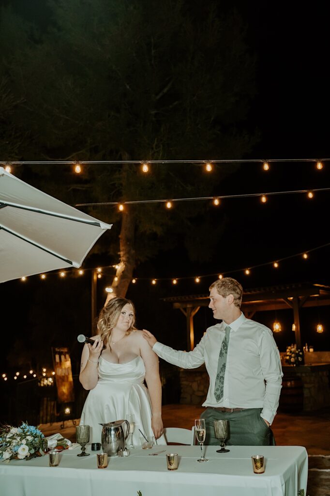 A bride gives a toast at her wedding and stands looking at the camera while her husband looks lovingly over at her.  Their table has glass gold votives on it and there are string lights hanging behind them in the background.  