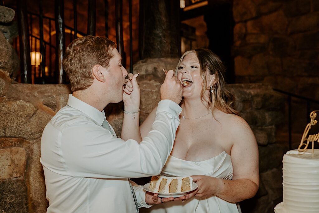 A bride and groom eat their wedding cake together on their wedding day.  The bride is wearing a scoop neck wedding dress with pleats along the side, her husband wears a plane white button up with a green tie. 