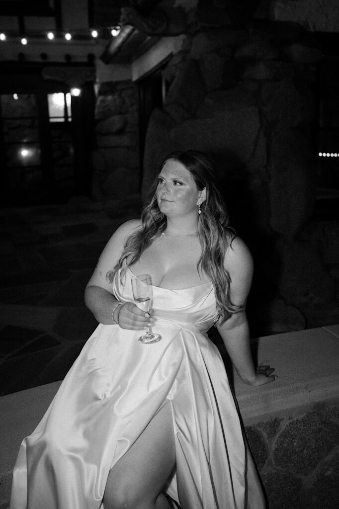 A bride sits on a rock wall at Mt Woodson Castle during her wedding reception in California sipping champagne. 