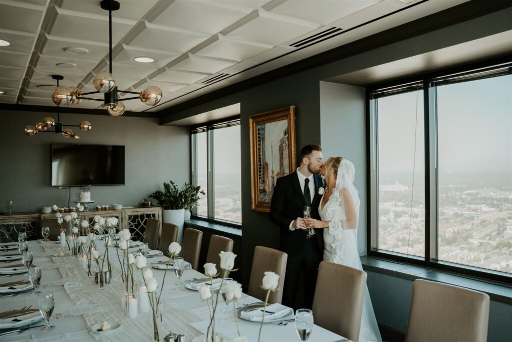 couple kissing while overlooking their great intimate wedding dinner table with white roses and candles in a skyscraper overlooking downtown indy 