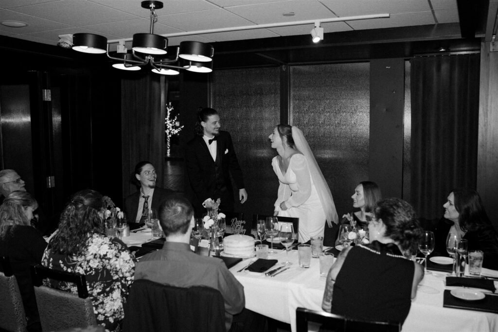 black and white photo of bride and groom laughing at one another surrounded by family at their great intimate wedding dinner with cake