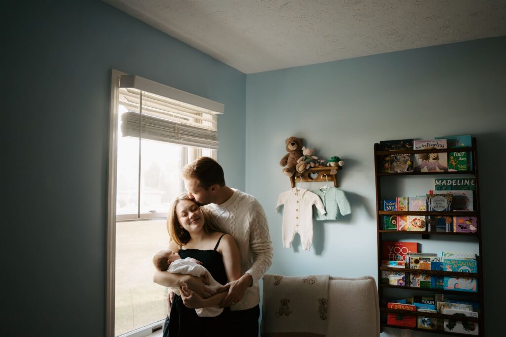 couple standing and kissing at window in blue nursery while holding their newborn
