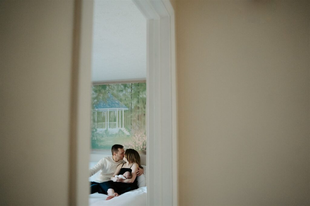 couple kissing while sitting in bed and holding newborn