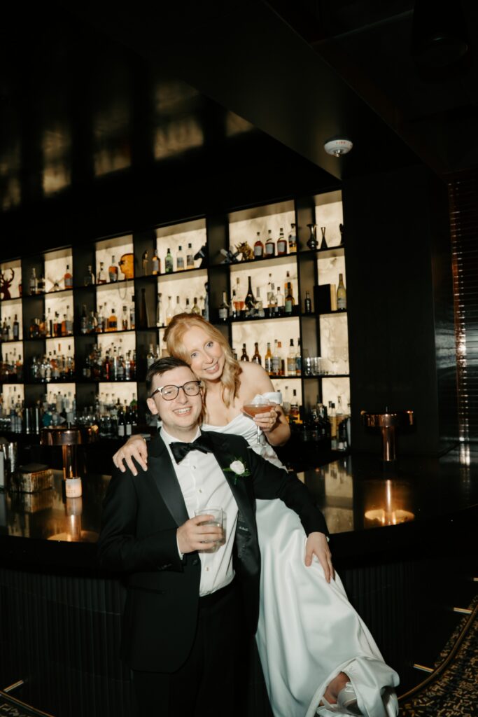couple laughing at camera while bride sits on bar next to groom